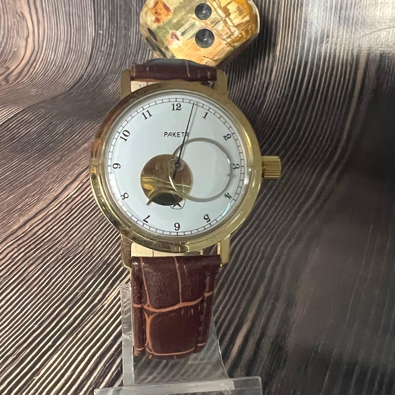 May include: A gold-toned wristwatch with a white face and brown leather band. The watch face features black numerals and the word "PAKETA". The band has a crocodile pattern. The watch is displayed on a clear stand.