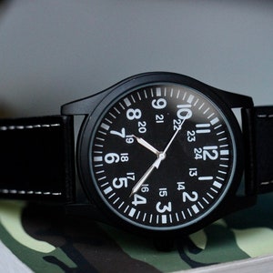 May include: Black wristwatch with a black face and white numerals. The watch has a black leather band with white stitching. The watch is on a camouflage pattern surface.