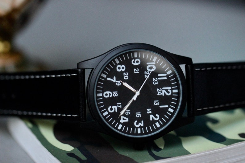 May include: Black wristwatch with a black face and white numerals. The watch has a black leather band with white stitching. The watch is on a camouflage pattern surface.
