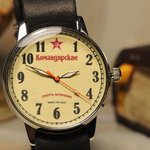 May include: A vintage wristwatch with a cream-colored face, black numerals, and red accents. The watch features a black leather band and a stainless steel case. The face includes Cyrillic text and a red star emblem. The watch is a classic timepiece.