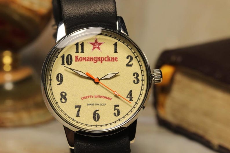 May include: A vintage wristwatch with a cream-colored face, black numerals, and red accents. The watch features a black leather band and a stainless steel case. The face includes Cyrillic text and a red star emblem. The watch is a classic timepiece.