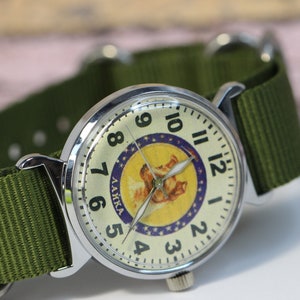 May include: A silver wristwatch with a white face and black numbers. The watch face features a yellow circle with a cartoon dog and the text "LAIKA" in Russian. The watch has a green nylon strap.