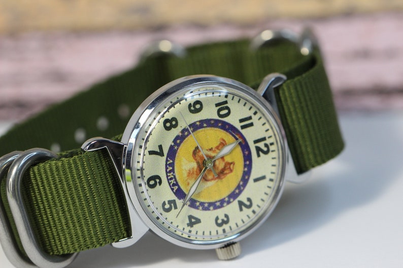May include: A silver wristwatch with a white face and black numbers. The watch face features a yellow circle with a cartoon dog and the text "LAIKA" in Russian. The watch has a green nylon strap.