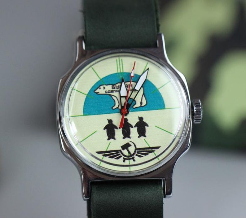 May include: A silver wristwatch with a green leather band. The watch face has a white background with a blue and green polar bear design and three penguins. The text "БУДЬ" and "СОСНЯ" is printed on the watch face.