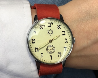 Puede incluir: Un reloj de pulsera plateado con una correa de cuero roja. La esfera del reloj tiene un fondo blanco con números hebreos negros y una estrella de seis puntas en el centro.