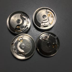 Puede incluir: Cuatro movimientos de reloj desmontados, que muestran intrincados mecanismos internos. Los componentes plateados incluyen engranajes, resortes y otras piezas peque&ntilde;as, dispuestas en una configuraci&oacute;n circular. La imagen est&aacute; tomada desde una perspectiva cenital, destacando la detallada artesan&iacute;a.