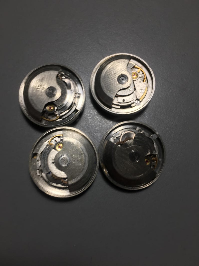 Puede incluir: Cuatro movimientos de reloj desmontados, que muestran intrincados mecanismos internos. Los componentes plateados incluyen engranajes, resortes y otras piezas peque&ntilde;as, dispuestas en una configuraci&oacute;n circular. La imagen est&aacute; tomada desde una perspectiva cenital, destacando la detallada artesan&iacute;a.