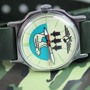May include: A silver wristwatch with a green leather band. The watch face is white with a blue and green design featuring a polar bear and three penguins. The text "БУРАН" is printed on the watch face.