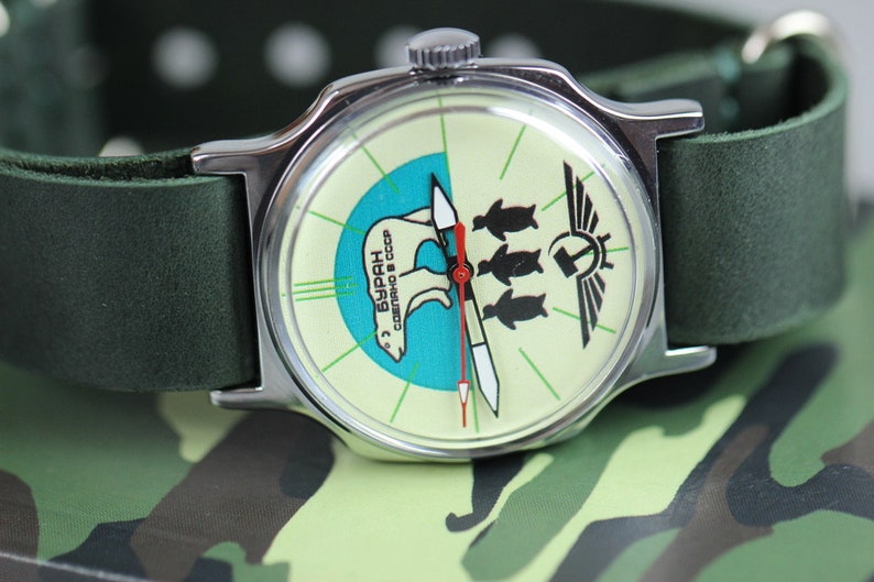 May include: A silver wristwatch with a green leather band. The watch face is white with a blue and green design featuring a polar bear and three penguins. The text "БУРАН" is printed on the watch face.