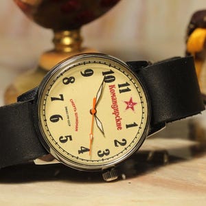 May include: Vintage wristwatch with a cream-colored face, black leather band, and silver-tone case. The watch face features black numerals, red text, and a red star. The watch is displayed on a light-colored surface.