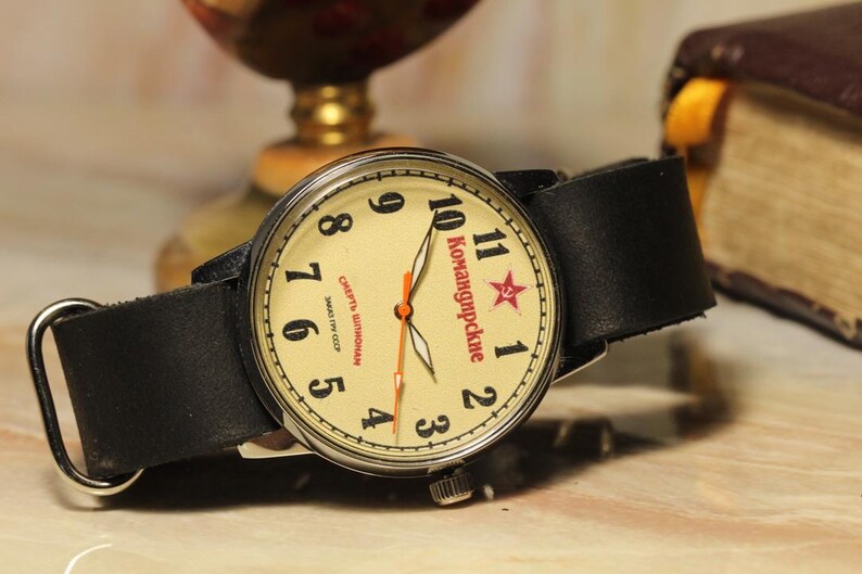 May include: Vintage wristwatch with a cream-colored face, black leather band, and silver-tone case. The watch face features black numerals, red text, and a red star. The watch is displayed on a light-colored surface.
