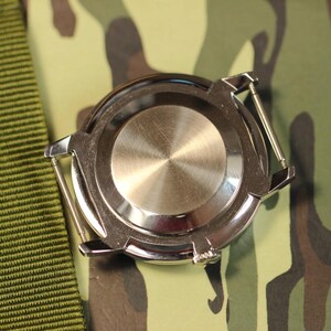May include: A silver watch with a round, stainless steel case back. The watch is on a green camouflage fabric background.