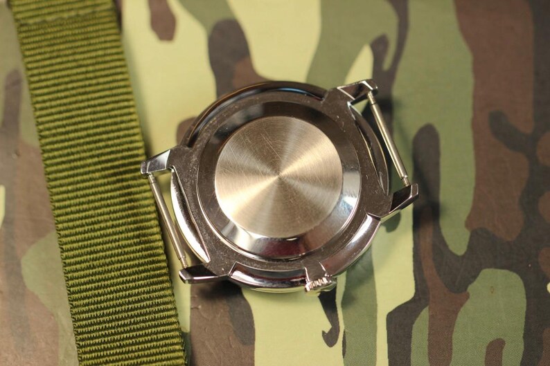 May include: A silver watch with a round, stainless steel case back. The watch is on a green camouflage fabric background.