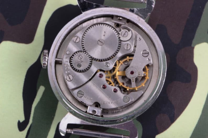 May include: Close-up view of the back of a silver wristwatch with a mechanical movement. The watch face is exposed, revealing the intricate gears and springs. The watch has a black and green camouflage background.