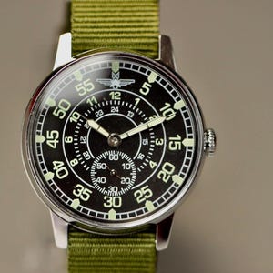 May include: A silver wristwatch with a black face and white markings. The watch has a green canvas band and a small airplane logo on the face.