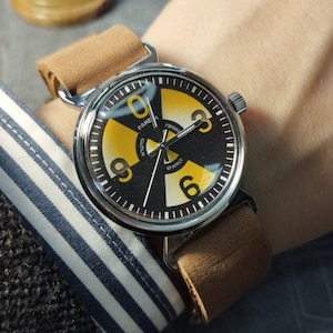 May include: A wristwatch featuring a black and yellow radiation symbol design. The watch has a silver case and a brown leather strap. The watch face displays the text "CAUTION" and "CONTACT".