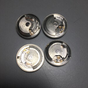 Puede incluir: Cuatro movimientos de reloj desmontados, que muestran intrincados componentes mec&aacute;nicos. Las piezas plateadas incluyen engranajes, resortes y otros elementos peque&ntilde;os, dispuestos dentro de marcos circulares. La imagen destaca la ingenier&iacute;a de precisi&oacute;n de los mecanismos del reloj.