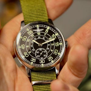 May include: A black and white analog wristwatch with a green canvas strap. The watch face has a large outer ring with numbers 5, 10, 15, 20, 25, 30, 35, 40, 45, 50, 55, and a smaller inner ring with numbers 1 through 12. The watch has a small second hand dial in the center.