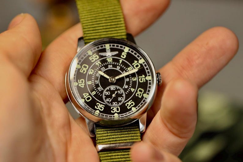 May include: A black and white analog wristwatch with a green canvas strap. The watch face has a large outer ring with numbers 5, 10, 15, 20, 25, 30, 35, 40, 45, 50, 55, and a smaller inner ring with numbers 1 through 12. The watch has a small second hand dial in the center.