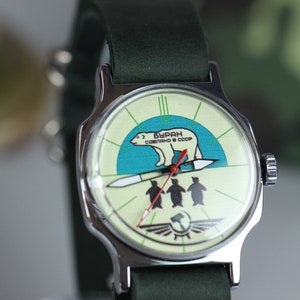 May include: A silver wristwatch with a green leather band. The watch face has a white background with a blue and green illustration of a polar bear walking on a rocket with three penguins below. The text on the watch face reads "БУРАН СОВЕТСКОГО СССР".