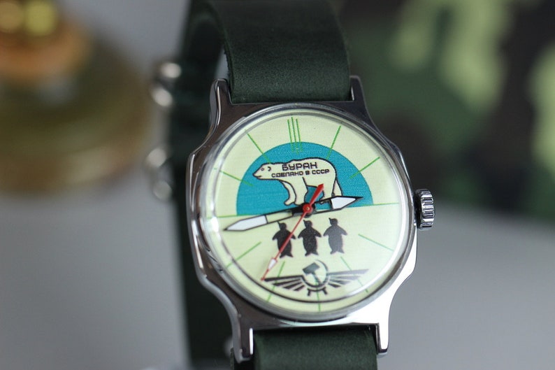 May include: A silver wristwatch with a green leather band. The watch face has a white background with a blue and green illustration of a polar bear walking on a rocket with three penguins below. The text on the watch face reads "БУРАН СОВЕТСКОГО СССР".
