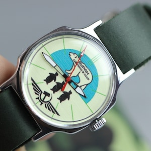 May include: A vintage wristwatch with a green leather band. The watch face is white with a green and blue design featuring a polar bear and penguins. The text "СССР" and "ЧАСЫ" is printed on the watch face.