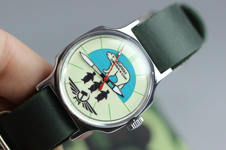 May include: A vintage wristwatch with a green leather band. The watch face is white with a green and blue design featuring a polar bear and penguins. The text "СССР" and "ЧАСЫ" is printed on the watch face.