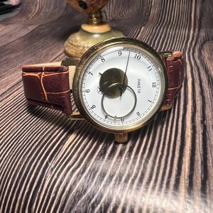May include: A vintage gold-toned wristwatch with a white face, black numerals, and a brown leather band. The watch features a unique subdial and is set against a wooden background. The watch is a classic timepiece.