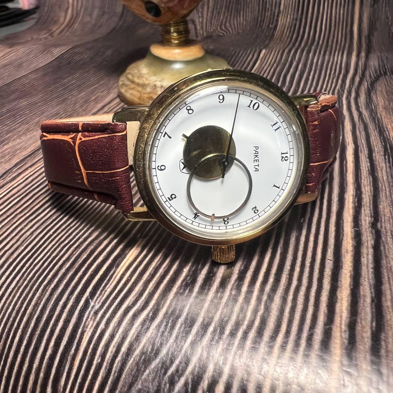 May include: A vintage gold-toned wristwatch with a white face, black numerals, and a brown leather band. The watch features a unique subdial and is set against a wooden background. The watch is a classic timepiece.