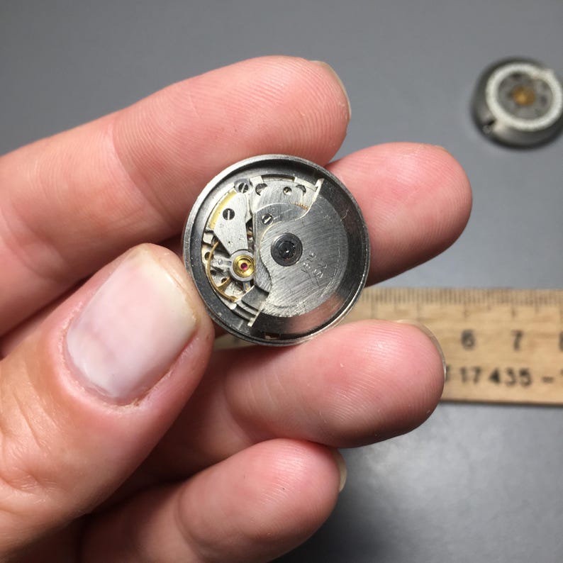 Puede incluir: Primer plano de un movimiento de reloj, mostrando engranajes, muelles y volante. Los componentes met&aacute;licos son plateados y dorados, con un peque&ntilde;o tornillo negro en el centro. Otro componente del reloj es visible en el fondo.