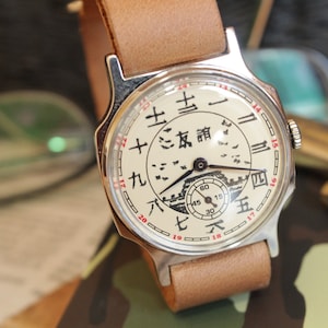 May include: A vintage wristwatch with a cream-colored face featuring Chinese characters and a small seconds dial. The watch has a silver-toned case and a light brown leather strap. The watch face also has a drawing of a building.