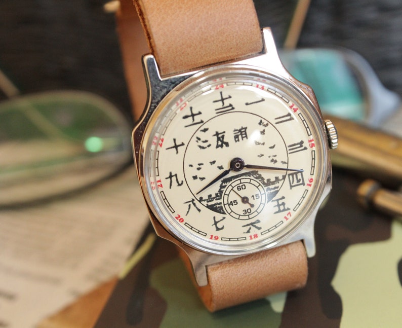 May include: A vintage wristwatch with a cream-colored face featuring Chinese characters and a small seconds dial. The watch has a silver-toned case and a light brown leather strap. The watch face also has a drawing of a building.