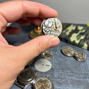 以下が含まれることがあります： 指の間に挟まれたヴィンテージ時計のムーブメントのクローズアップ。複雑な歯車とコンポーネントが映し出されています。時計の文字盤はシルバーとゴールドで、複雑な機械的デザインです。他の時計部品と時計が背景に見えます。