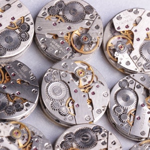 May include: Close-up of multiple silver watch movements with intricate gears and mechanisms. The watch movements are arranged in a circular pattern on a white surface.