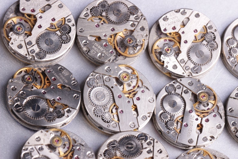 May include: Close-up of multiple silver watch movements with intricate gears and mechanisms. The watch movements are arranged in a circular pattern on a white surface.