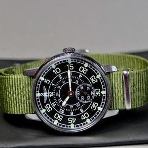 May include: A black and silver wristwatch with a green nylon strap. The watch face has a black background with white numbers and markings. The watch has a second hand and a small subdial.