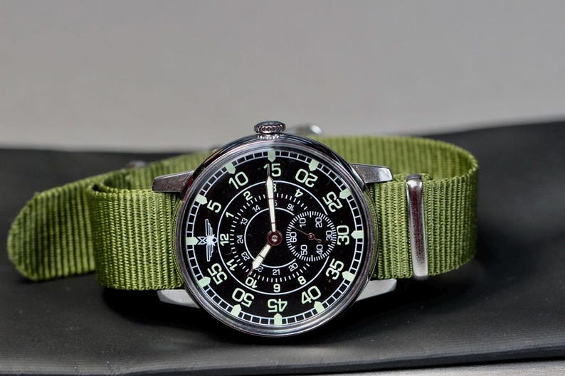 May include: A black and silver wristwatch with a green nylon strap. The watch face has a black background with white numbers and markings. The watch has a second hand and a small subdial.