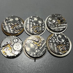 以下が含まれることがあります： 露出した歯車と複雑な機械部品を備えた、6つのヴィンテージ時計ムーブメント。時計部品は、金色の歯車と赤い宝石を備えた銀色の金属が特徴です。ムーブメントは円形で、分解の程度が異なります。