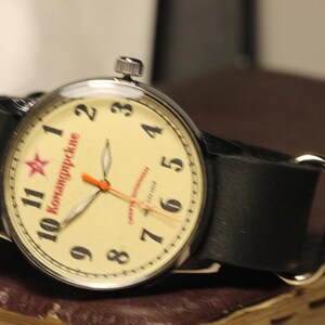 May include: A vintage wristwatch with a cream-colored face, black leather band, and silver-tone case. The watch face features black numerals, red text, and a red star. The hands are orange.