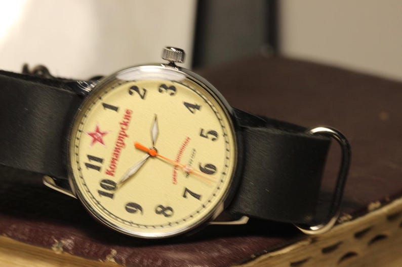 May include: A vintage wristwatch with a cream-colored face, black leather band, and silver-tone case. The watch face features black numerals, red text, and a red star. The hands are orange.