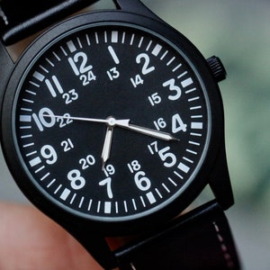 May include: Close-up of a black wristwatch with a black face and white numerals. The watch has a black leather band and a black case. The watch face displays both 12-hour and 24-hour time. The hands are white.
