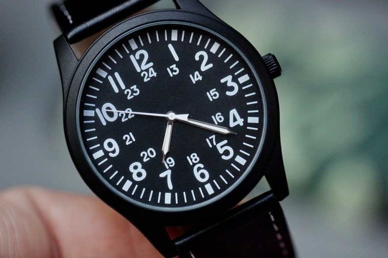 May include: Close-up of a black wristwatch with a black face and white numerals. The watch has a black leather band and a black case. The watch face displays both 12-hour and 24-hour time. The hands are white.