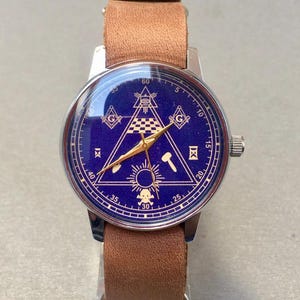 May include: A wristwatch with a blue face featuring gold-colored Masonic symbols, including a compass, square, and all-seeing eye. The watch has a silver-colored case and a brown leather band.