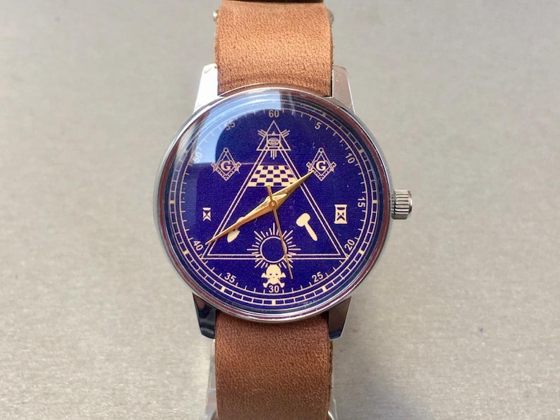 May include: A wristwatch with a blue face featuring gold-colored Masonic symbols, including a compass, square, and all-seeing eye. The watch has a silver-colored case and a brown leather band.