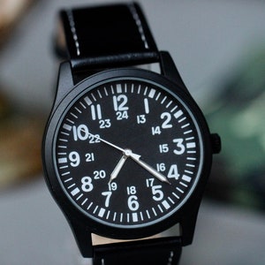 May include: Black wristwatch with a black leather band and white stitching. The watch face is black with white numerals and hands. The watch has a 24-hour clock face. The watch case is black.