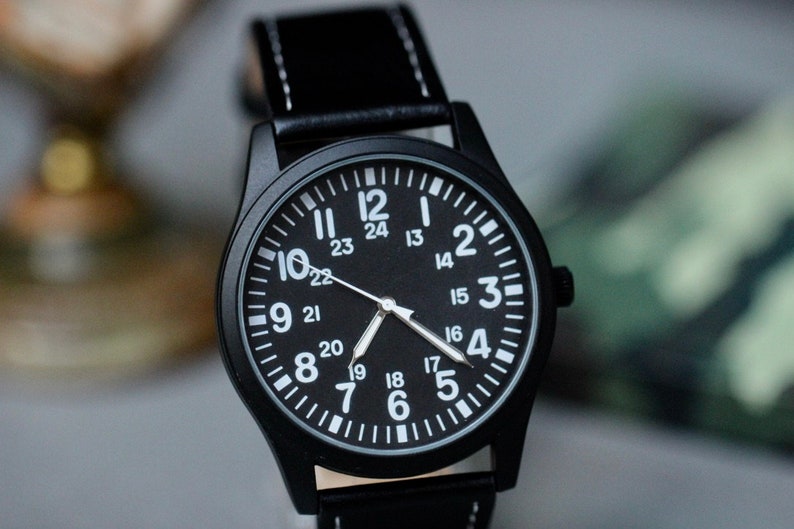 May include: Black wristwatch with a black leather band and white stitching. The watch face is black with white numerals and hands. The watch has a 24-hour clock face. The watch case is black.