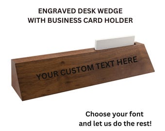 Puede incluir: Una cuña de escritorio de madera marrón con un tarjetero. La cuña tiene el texto "YOUR CUSTOM TEXT HERE" grabado en la parte delantera. Una tarjeta de visita blanca está en el soporte. El texto "ENGRAVED DESK WEDGE WITH BUSINESS CARD HOLDER" está en la parte superior.