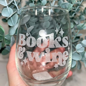 Op de afbeelding: Een helder, stamloos wijnglas met de woorden "Books & Wine" in een retro lettertype. Het glas heeft ster- en diamantaccenten. Het glas wordt vastgehouden voor een achtergrond van groen gebladerte.