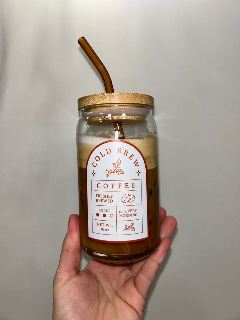 May include: A glass jar with a bamboo lid and a brown glass straw filled with cold brew coffee. The jar has a label that reads "Cold Brew + Coffee + Freshly Brewed Roast O Est. Every Morning Net Wt. 16 oz"