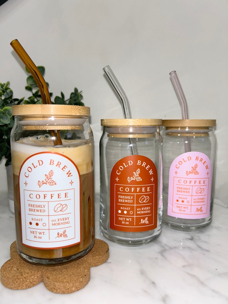 May include: Three glass jars with bamboo lids and glass straws. Each jar has a label that says "Cold Brew Coffee" with details about the coffee, including "Freshly Brewed", "Roast", and "Est. Every Morning". The jars are on a white marble surface.